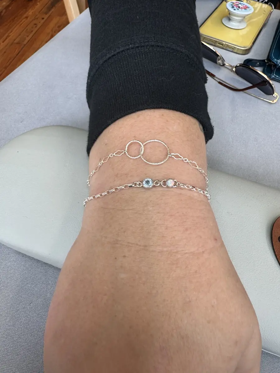 View of a person's leg resting on a book or cushion, with two silver anklets around their ankle. The background shows a table with sunglasses, a yellow tray, and a small bowl containing decorative beads or stones.