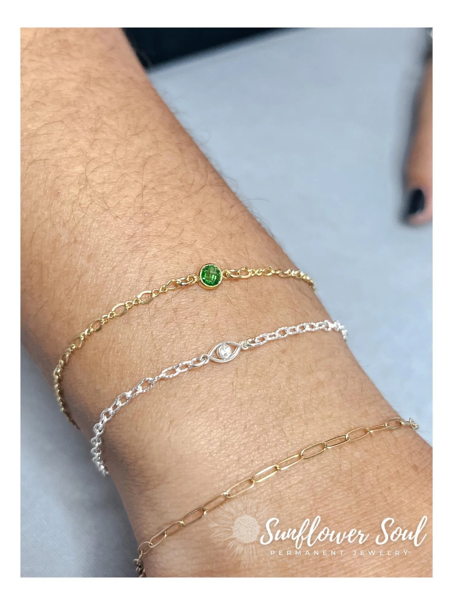 Four delicate bracelets on a person's wrist, each with different charms and colors, including a green stone, a clear stone, and plain gold and silver links, with a blurred background and Sunflower Soul Permanent Jewelry logo in the corner.