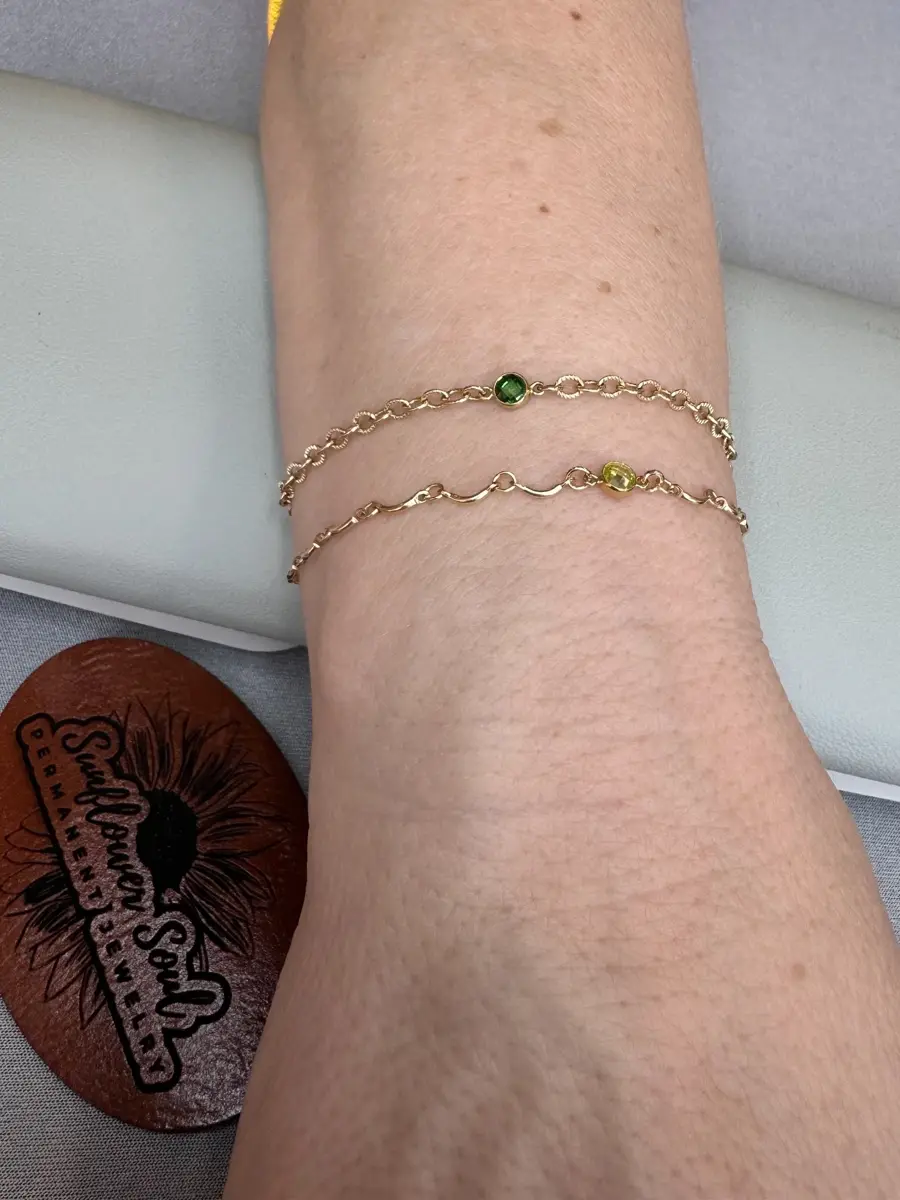 A person's forearm with two delicate gold chain bracelets, each with small green or yellow gemstone accents, resting on a gray surface with a leather circle that has branding.