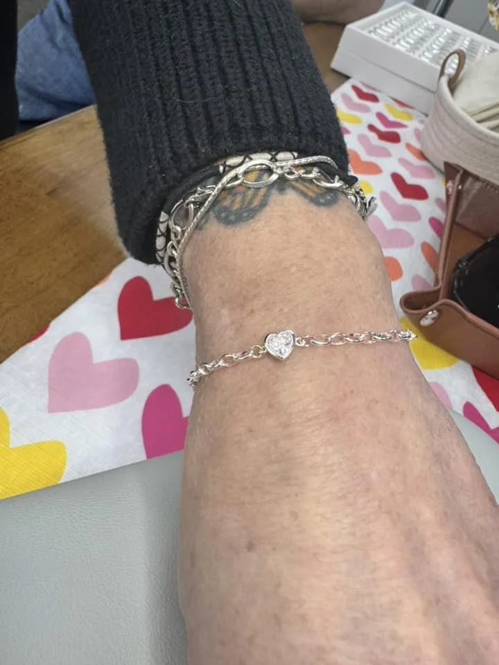 Close-up of a person's leg with several silver bracelets and an anklet, on a table with a pink and multicolored heart-patterned cloth.