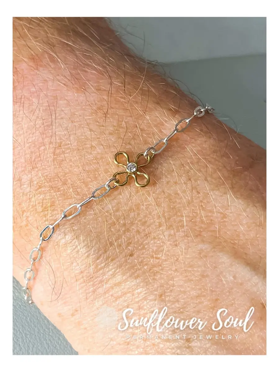 A close-up of a silver chain bracelet with linked oval loops, featuring a gold flower charm with a central gemstone, worn on a person's wrist with light-colored skin and fine hair. The background is plain and outdoors.