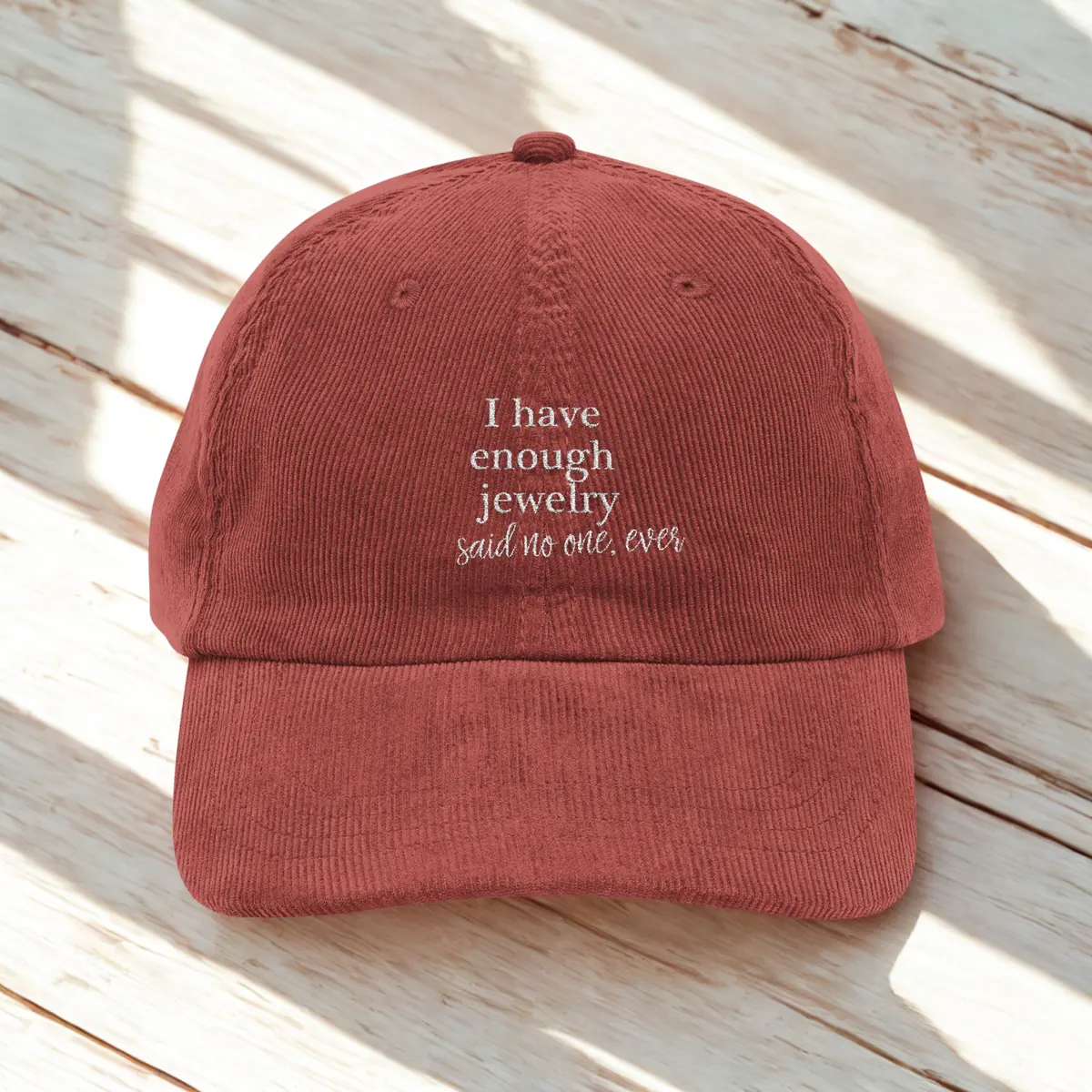 A rust-colored baseball cap with white embroidered text that reads, "I have enough jewelry said no one, ever," placed on a light wooden surface.