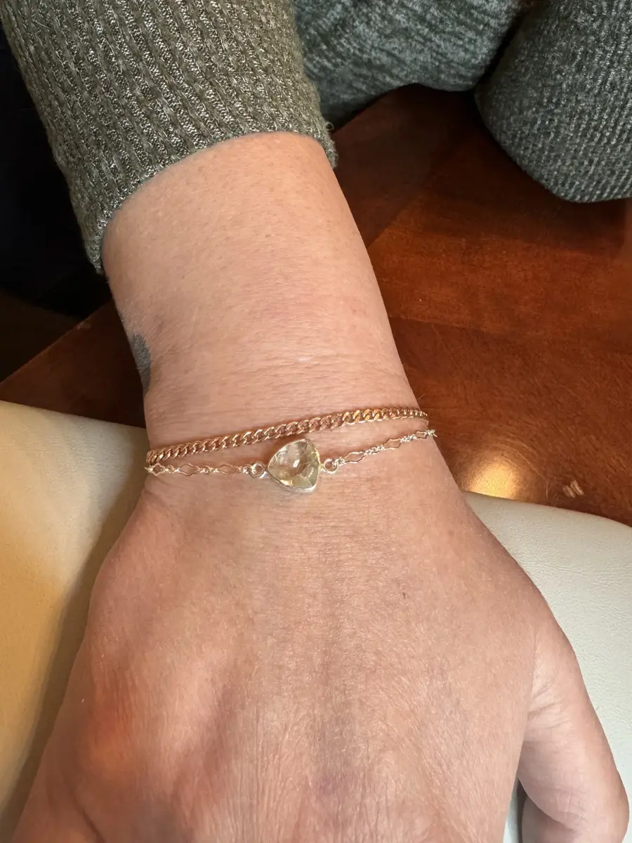 A person's arm resting on a wooden surface, wearing two bracelets with a heart-shaped charm and a textured sweater.