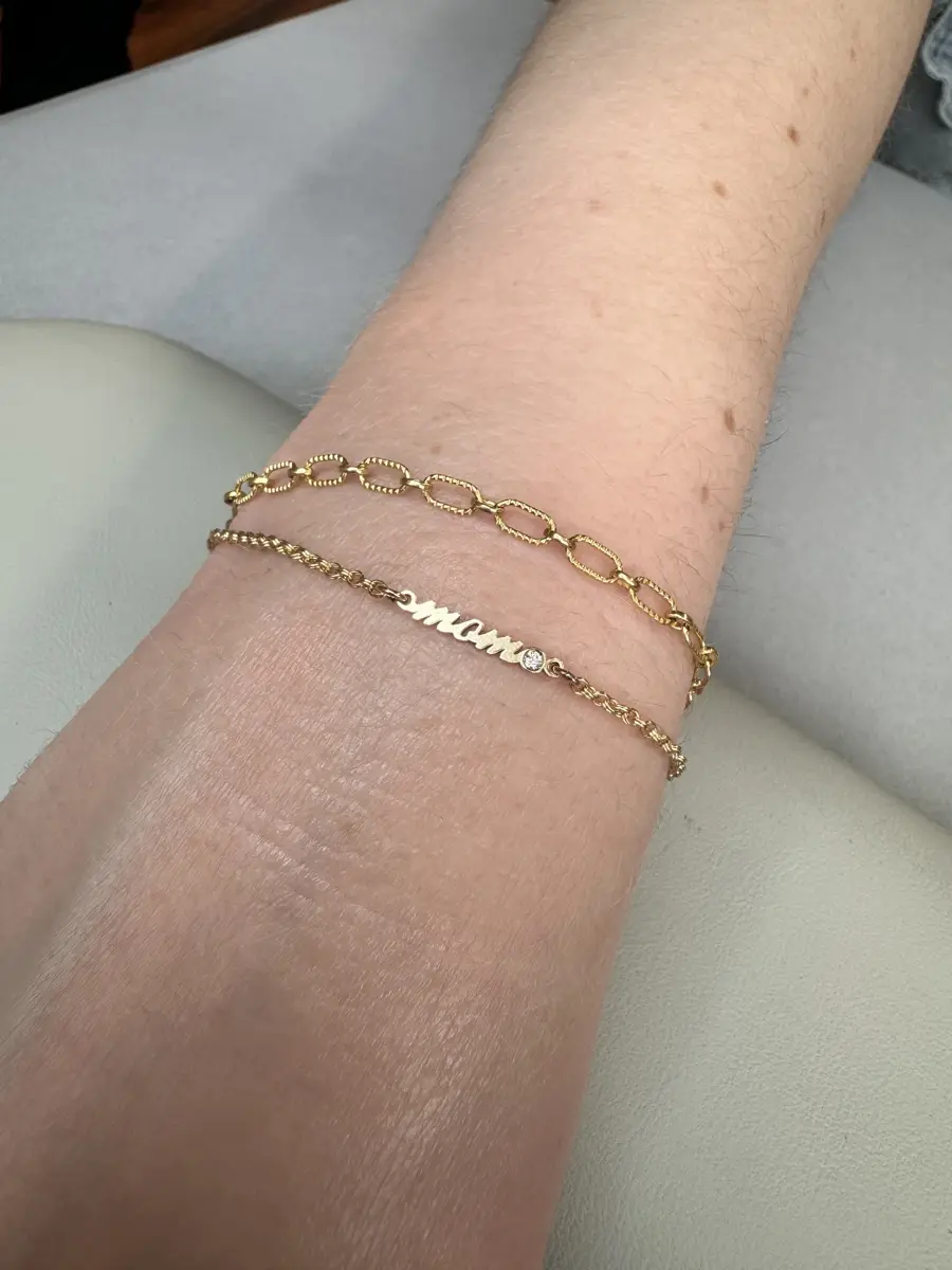 A person's wrist wearing three gold bracelets, one with a nameplate and a small gemstone, another with gold links, and the third with larger oval links. The background shows a light-colored surface and part of the person's arm with some freckles.