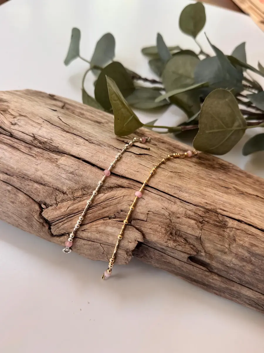 Two delicate beaded bracelets, one silver and one gold, resting on a piece of driftwood with eucalyptus leaves in the background.