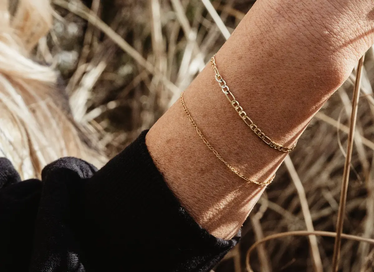 A person's wrist adorned with two gold bracelets, one with larger chain links and the other with smaller, delicate links, against a background of dried grass.