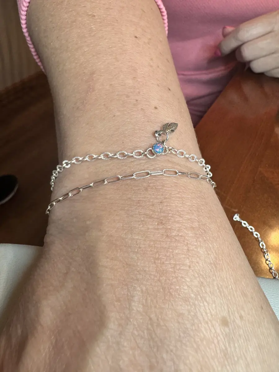 Close-up of a person's wrist with two silver bracelets, one with a small heart, a blue and pink charm, and a feather, resting on a wooden surface.