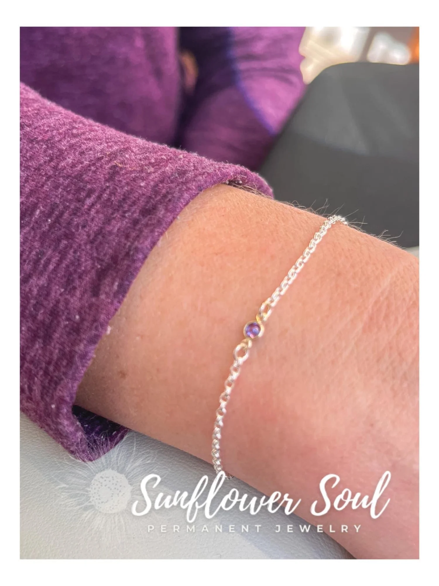 Close-up of a wrist wearing a delicate silver bracelet with a purple stone, resting on purple fabric, with text advertising Sunflower Soul Permanent Jewelry.