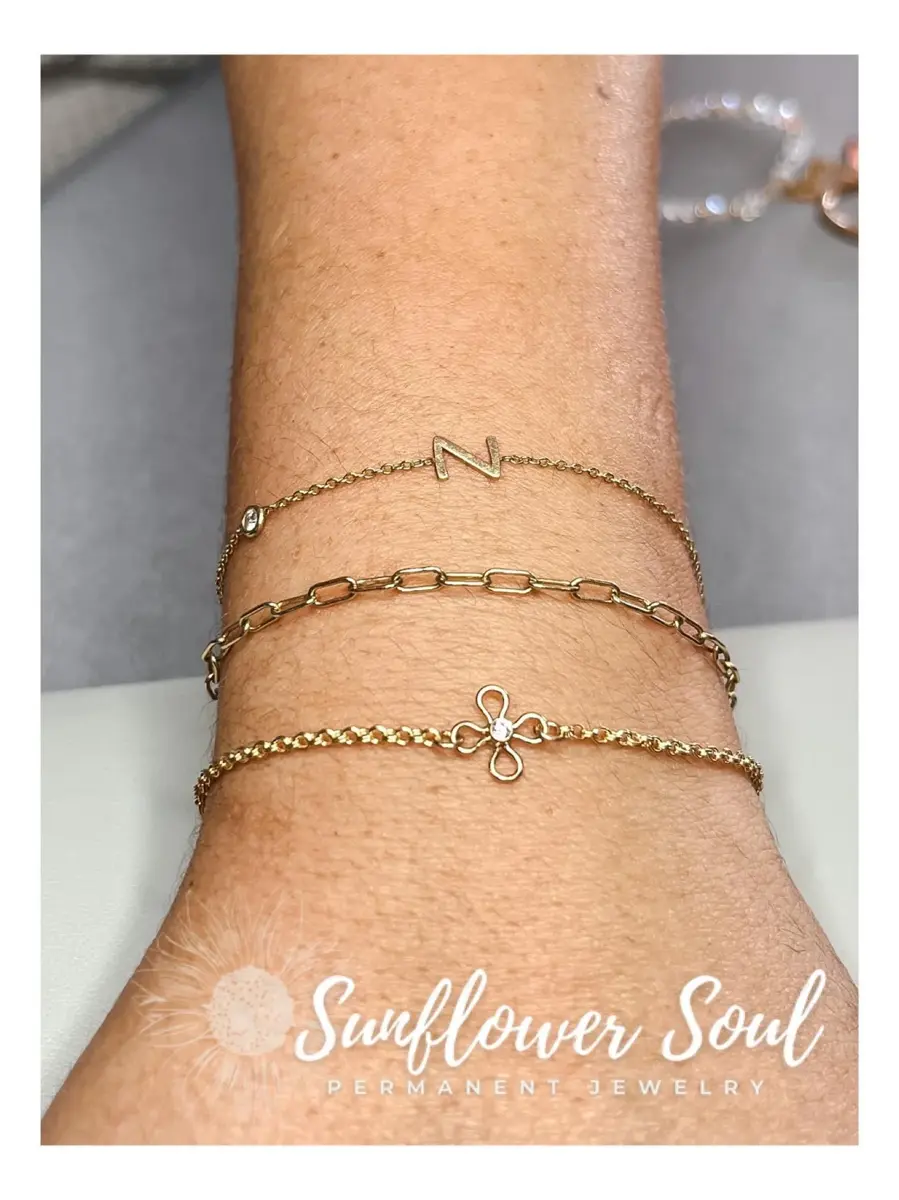 Three delicate gold bracelets on a wrist, each with different designs including a letter 'N', a chain link, and a flower with a small gemstone, with a logo for Sunflower Soul Permanent Jewelry at the bottom.
