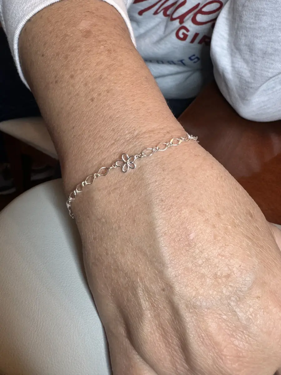 A person's wrist wearing a delicate silver bracelet with a small flower charm.