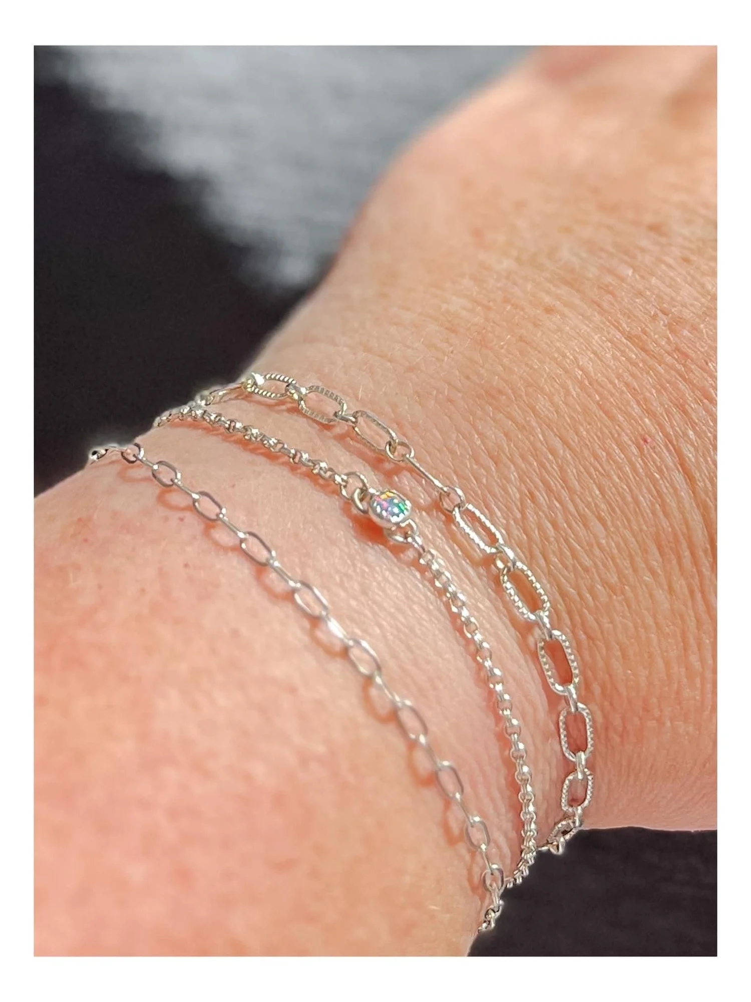 Close-up of a wrist with three silver chain bracelets, one with oval links, one with small beads, and one with a small iridescent charm, against a dark background.