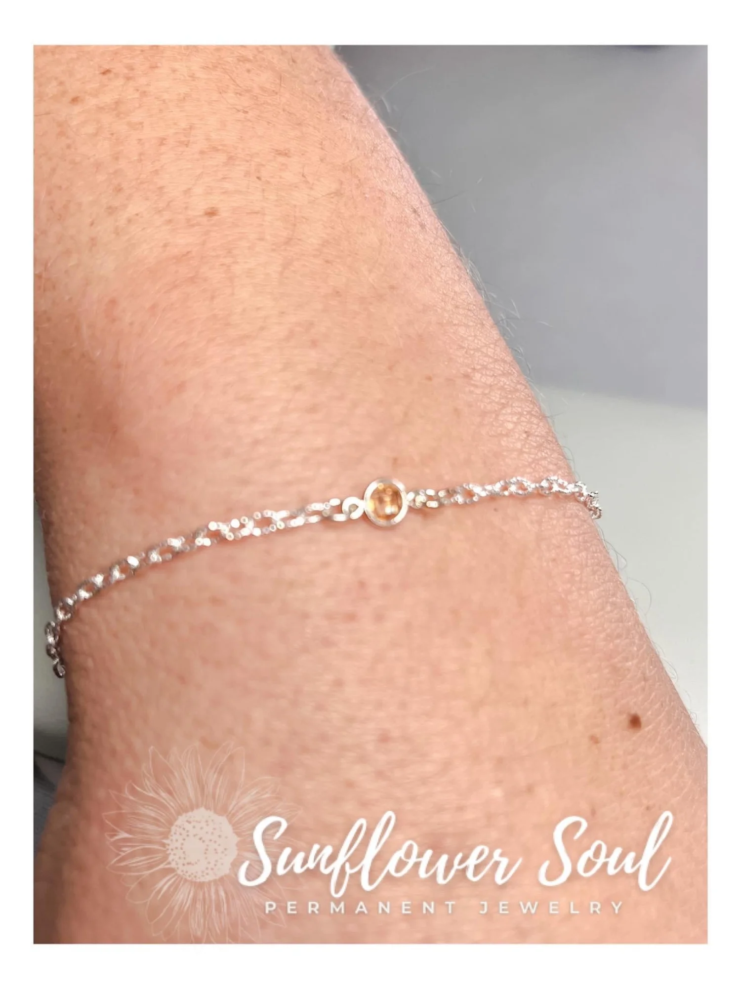 A close-up of a person's wrist with a delicate silver bracelet featuring small clear stones and a small round amber-colored gemstone in the center. The image includes a logo with a sunflower illustration and the text 'Sunflower Soul Permanent Jewelry