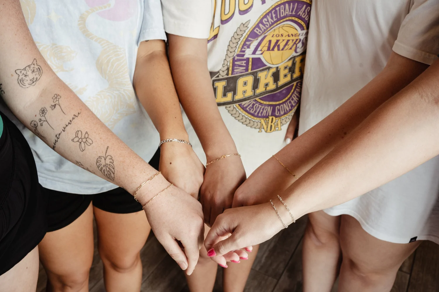 Four people with their hands stacked together in the center, wearing bracelets and casual clothing, including a t-shirt with a Lakers logo.
