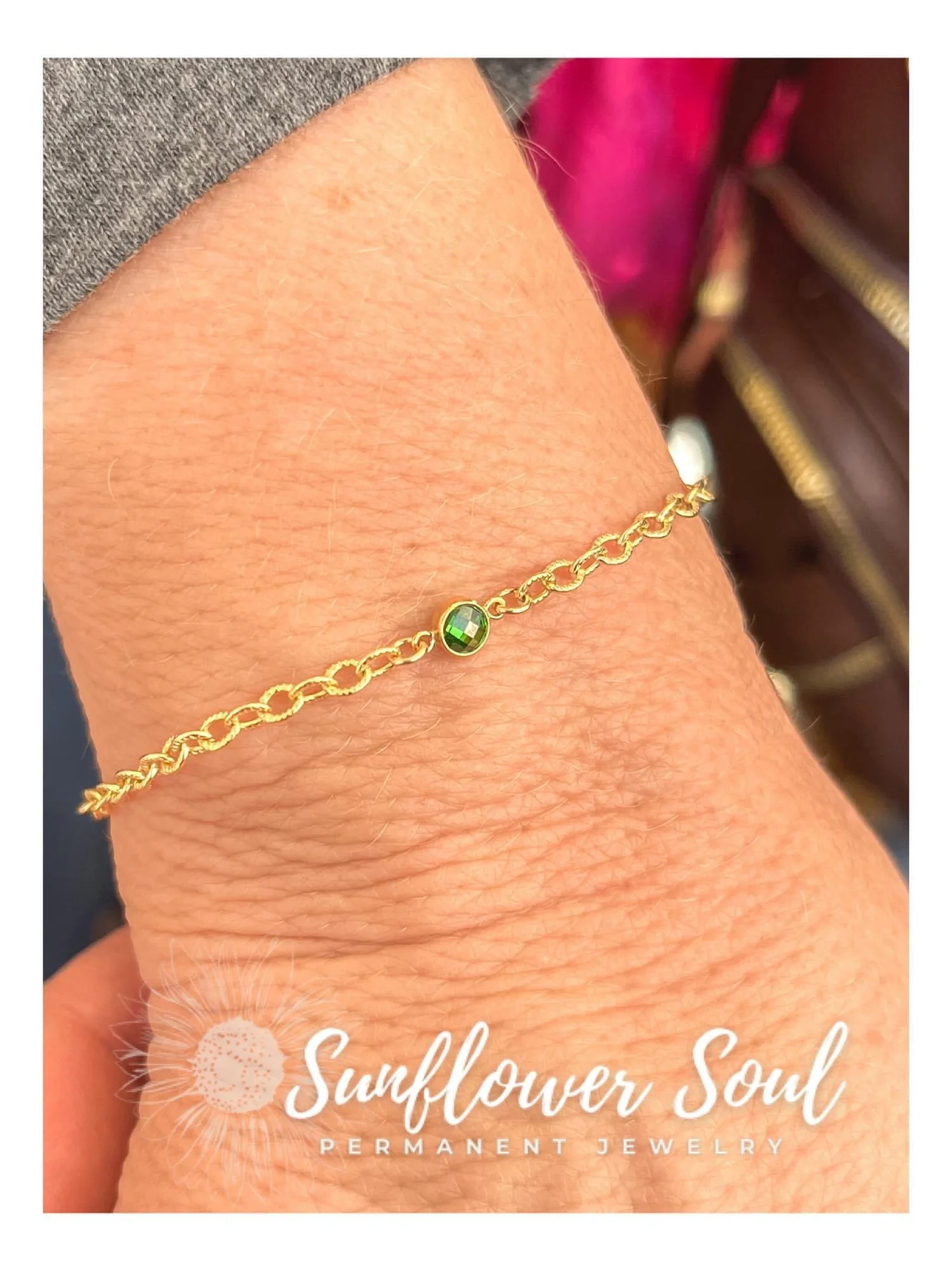 Close-up of a gold chain bracelet with a small green gemstone on a person's wrist, with Sunflower Soul Permanent Jewelry branding at the bottom.