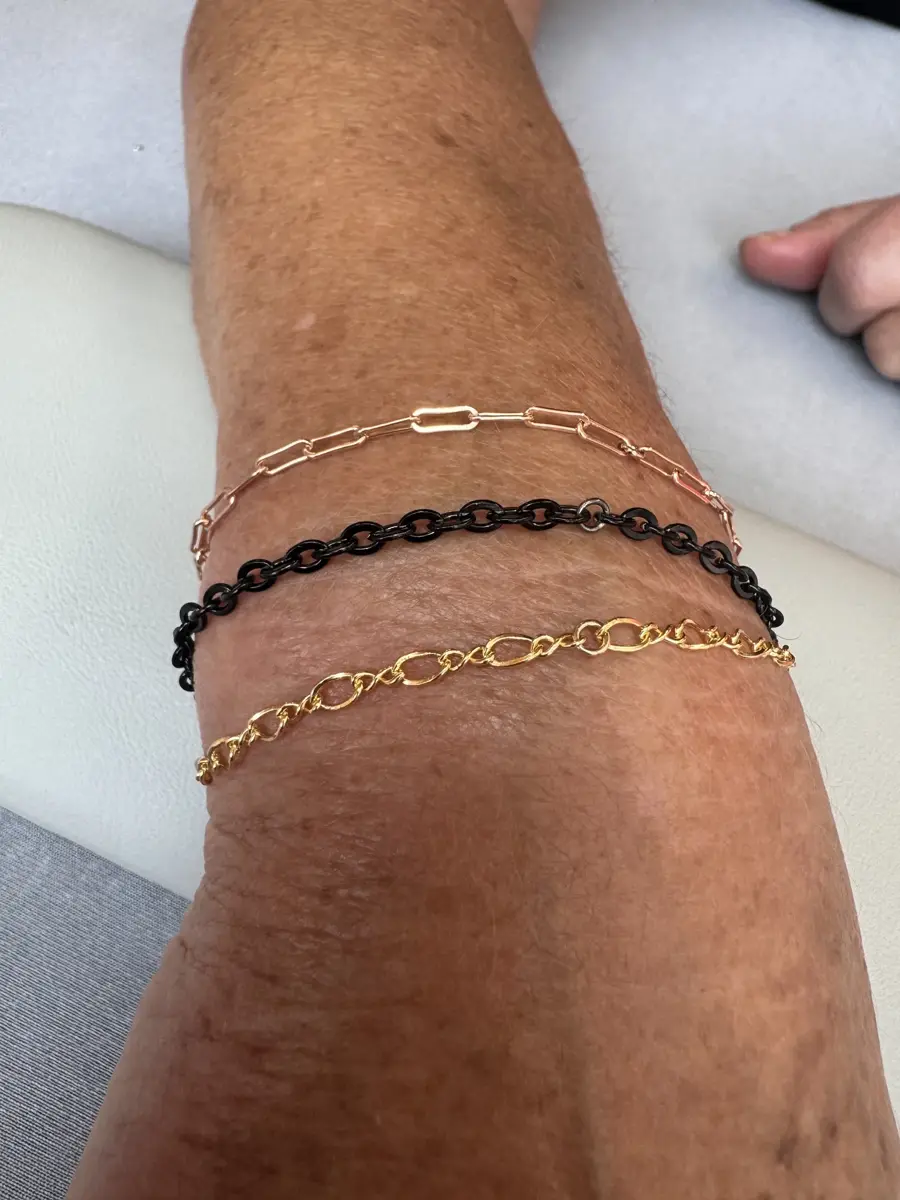 Three bracelets on a person's forearm: one black chain, one gold chain, and one rose gold chain.
