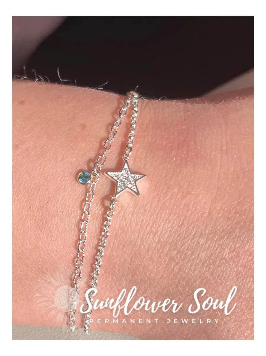 A close-up of a wrist with silver chain bracelets, one with a star-shaped pendant and the other with a small blue gem, against a black background. The image has the logo and text "Sunflower Soul Permanent Jewelry" at the bottom.