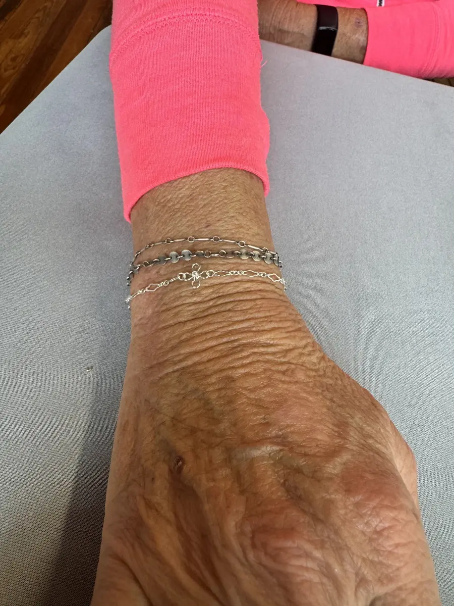 Close-up of an elderly person's wrist and hand resting on a gray surface, wearing multiple silver bracelets, with pink sleeve in the background.